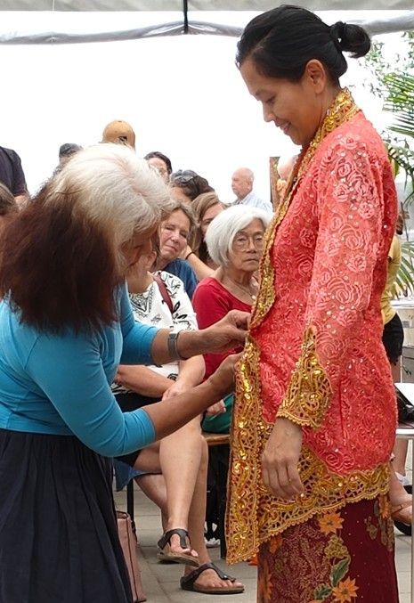 Een vrouw in een rode jurk ontvangt een bloemenkrans van de zittende aanwezigen tijdens een ceremonie in de openlucht.