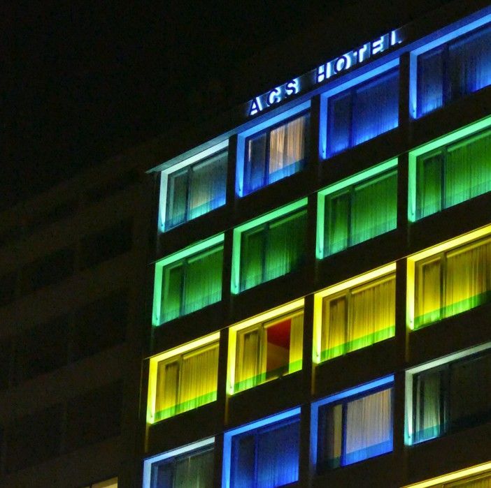 De gevel van het hotel 's nachts met verlichte ramen in groen, geel en blauw. Bovenaan staat de naam van het hotel: