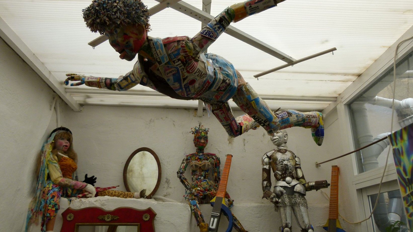 Een sculptuur van een persoon in de lucht, met uitgestrekte armen en benen, in een rommelige ruimte met andere sculpturen.