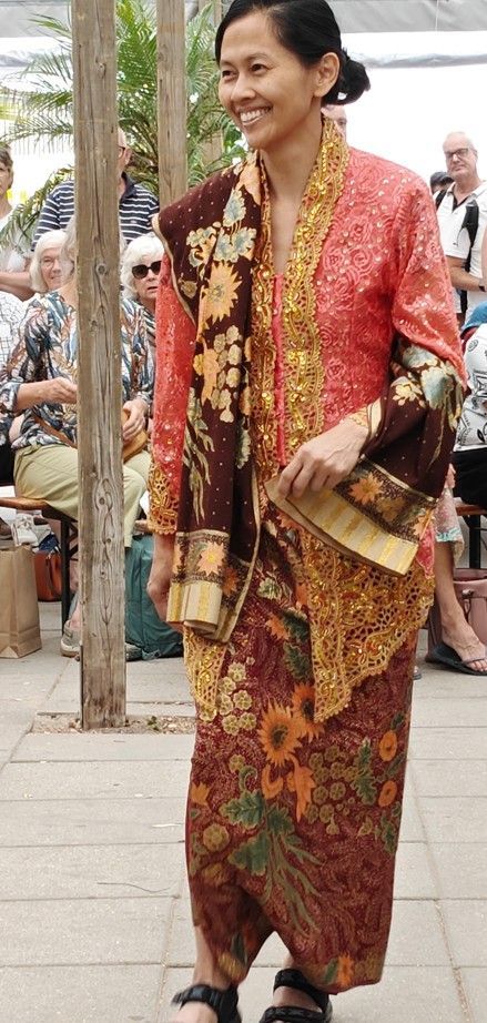 Vrouw in een traditionele rood-gouden jurk loopt buiten, lachend en met een sjaal in haar hand.