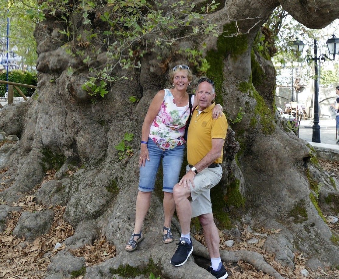 Een stel poseert naast een grote boom. De vrouw draagt ​​een top met bloemenprint, de man een geel shirt; beiden glimlachen. Buitenlocatie.