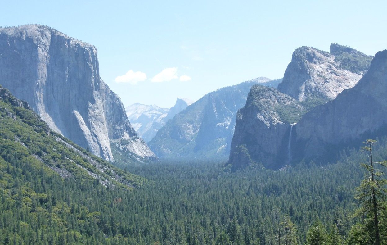 Uitzicht op Yosemite Valley met torenhoge granieten kliffen, weelderig bos en een waterval onder een blauwe hemel.