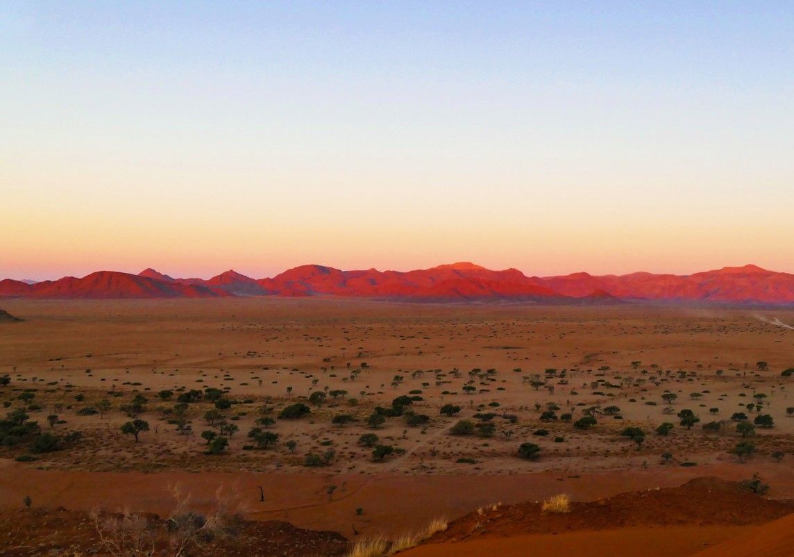 Woestijnlandschap bij zonsondergang, met in de verte rode bergen, een oranje-bruine ondergrond en een lichtblauwe lucht.