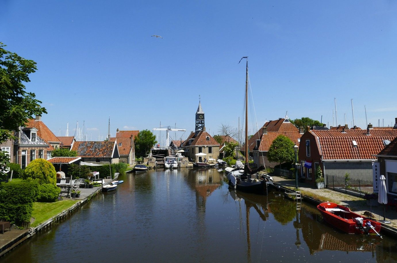 Een kanaal in een Nederlands dorp met boten, rode pannendaken, een blauwe lucht en een hoge toren.