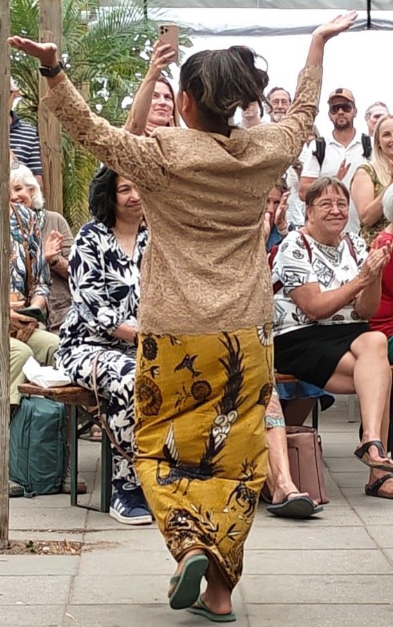 Een persoon danst met opgeheven armen in een beige top en een geel gestreepte rok voor een zittend publiek buiten.