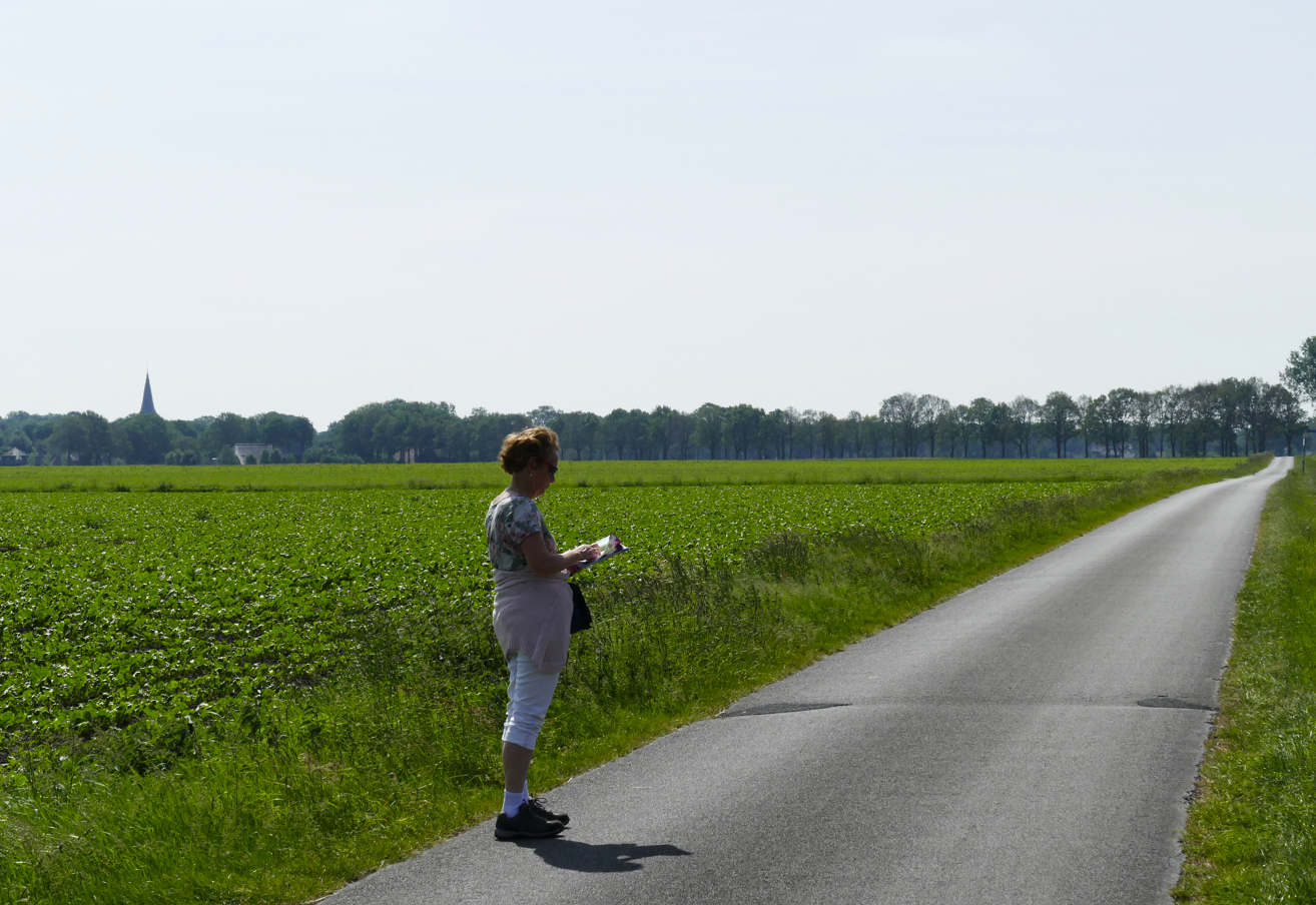 Een persoon staat langs een landweg en kijkt uit over een groen veld onder een heldere hemel.