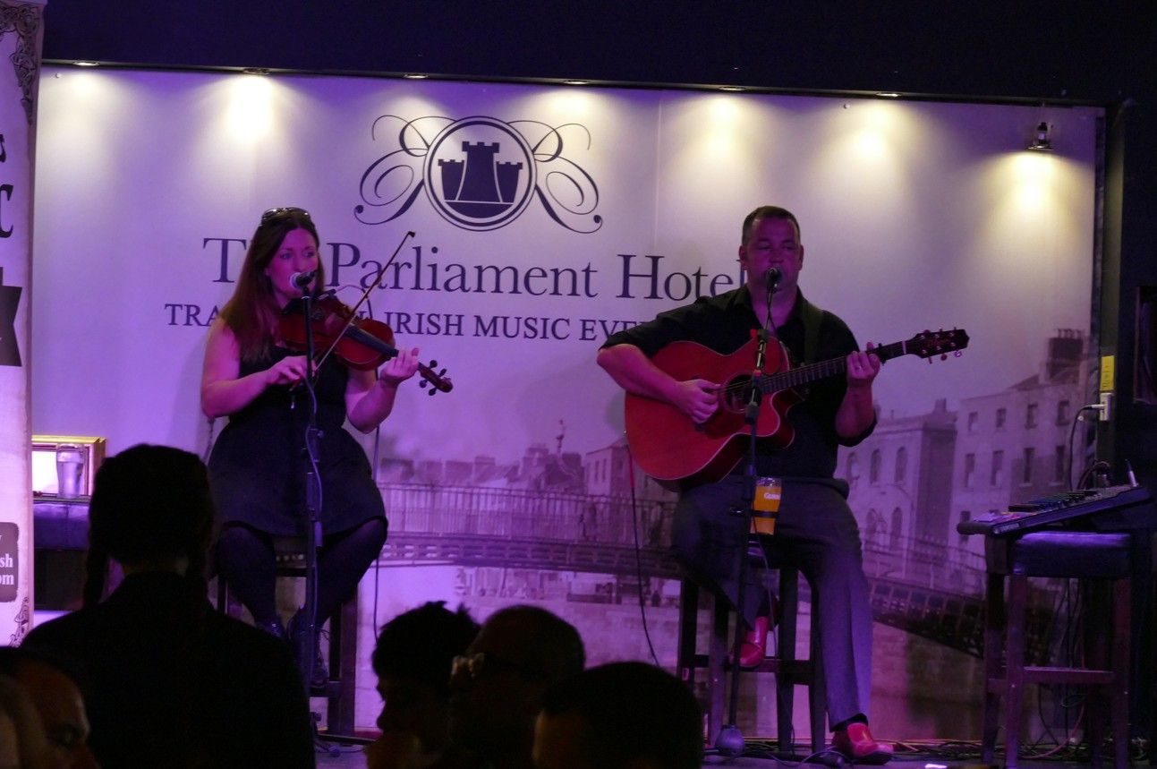 Muzikanten op het podium: vrouw met viool, man met gitaar, achtergrond: Parliament Hotel.