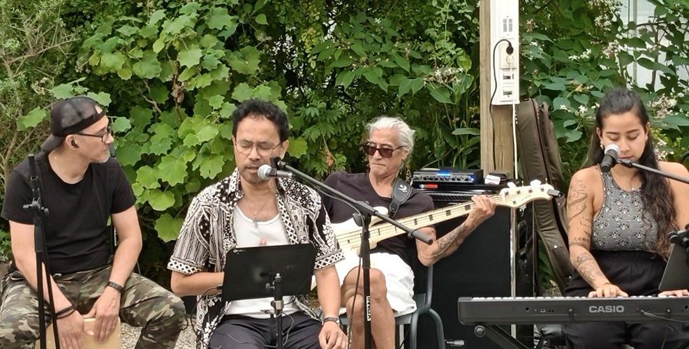Vier muzikanten treden buiten op, met microfoons, gitaar, keyboard en weelderig groen op de achtergrond.