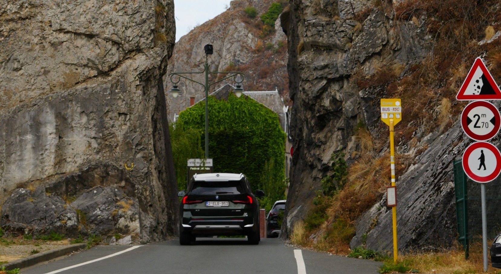 Een zwarte auto rijdt over een smalle, rotsachtige weg met aan de rechterkant een groene tunnel van heggen en verkeersborden.