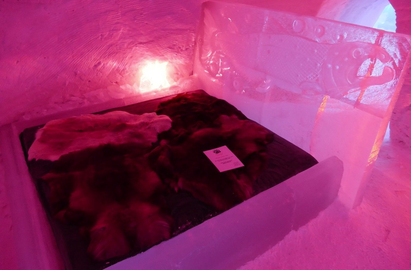 Een ijskleurig bedframe met donker beddengoed en een witte bontdeken, verlicht door roze licht.