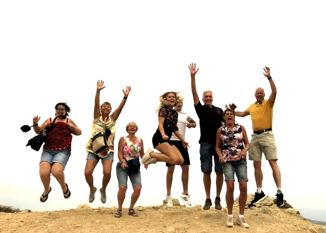 Een groep mensen springt op een aarden heuvel, met de armen omhoog. Bewolkte lucht.