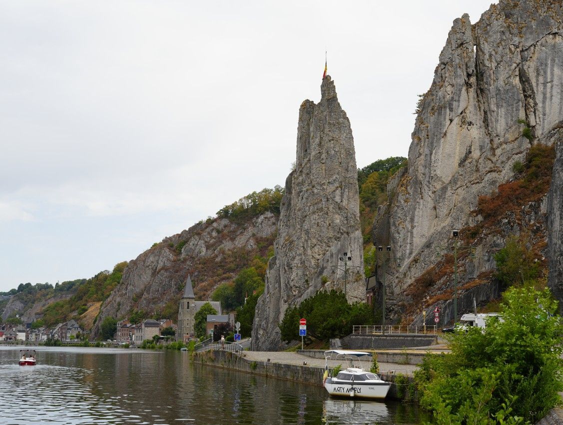 Een steile rotspiek langs een weg aan de rivier met aangemeerde boten en een dorpje op de klif onder een bewolkte hemel.
