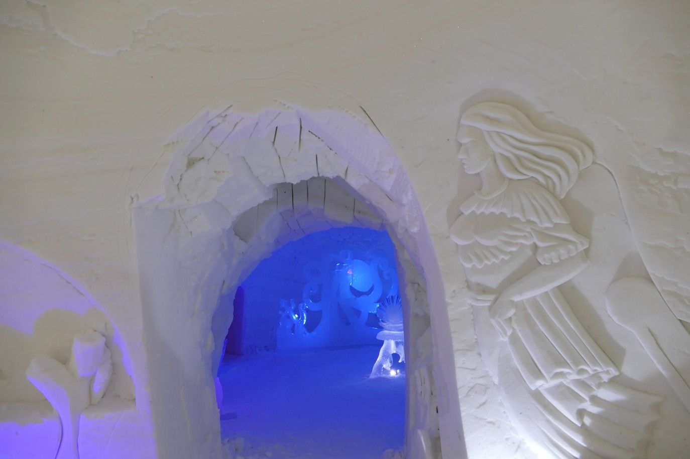 Interieur van een sneeuwsculptuur met een boog, blauw verlicht, en een gebeeldhouwde engelfiguur.