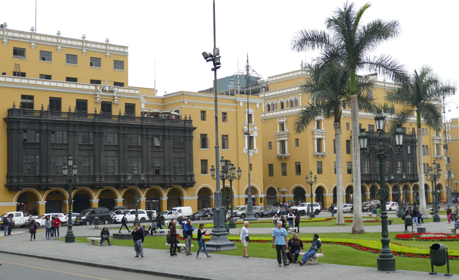 Plein in Lima, Peru, met gele gebouwen, palmbomen en mensen die wandelen en op bankjes zitten.