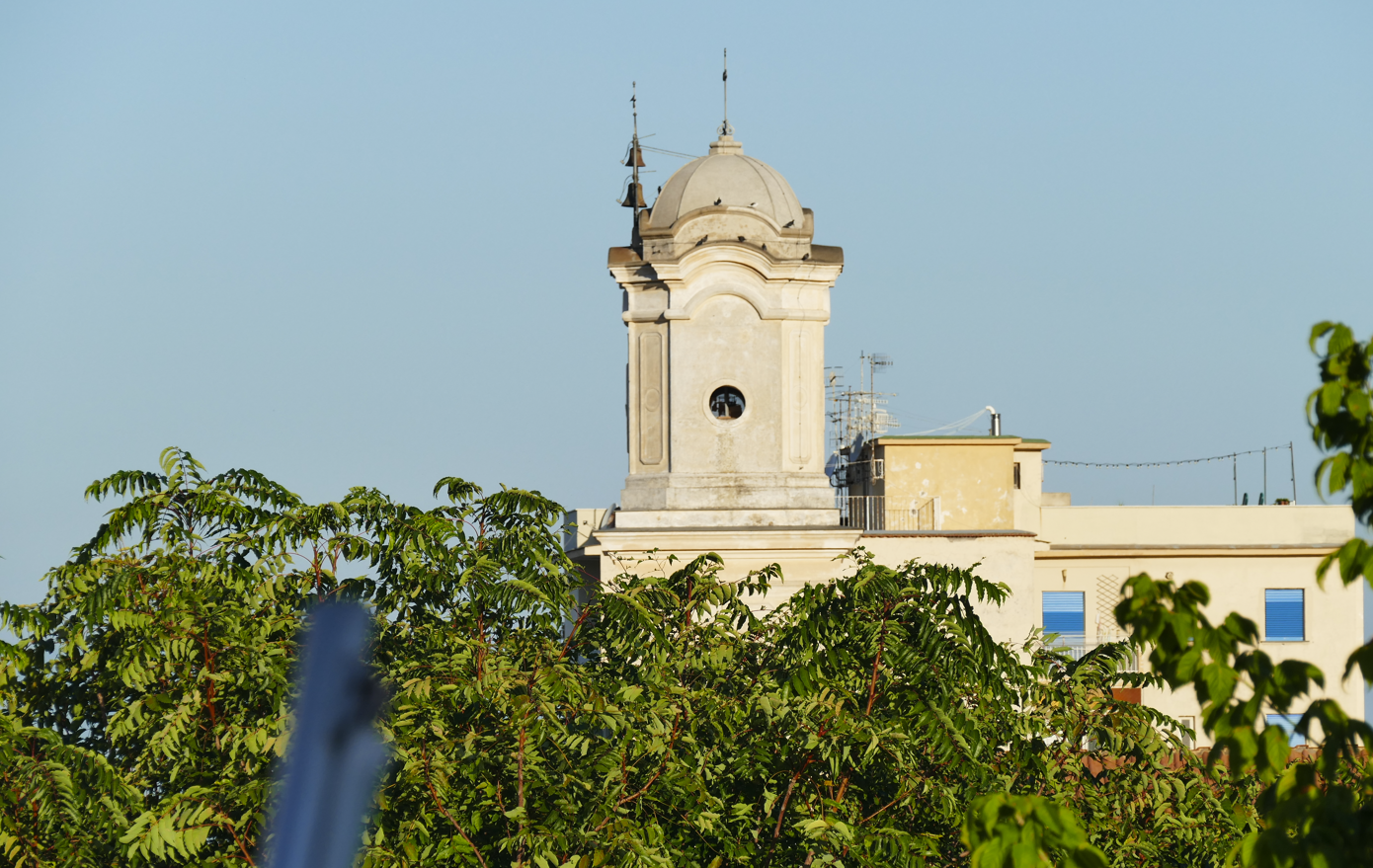 Een kerktoren met een koepel rijst boven groene bomen uit, onder een helderblauwe hemel.