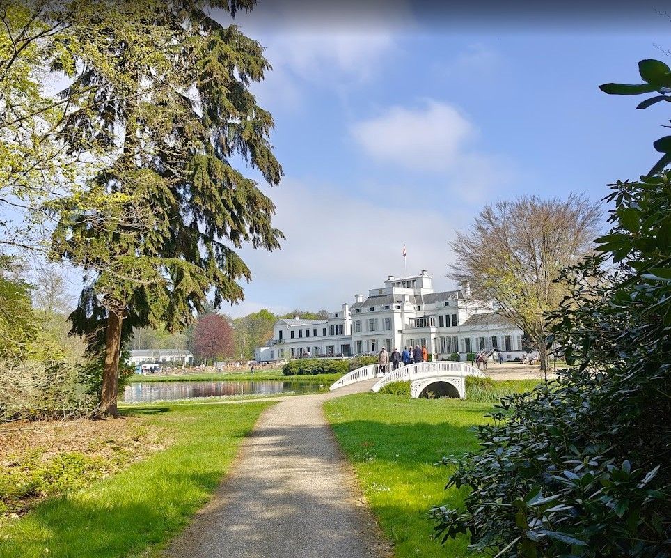 Het pad leidt naar een wit herenhuis met een brug over het water; het is een zonnige dag met bomen.