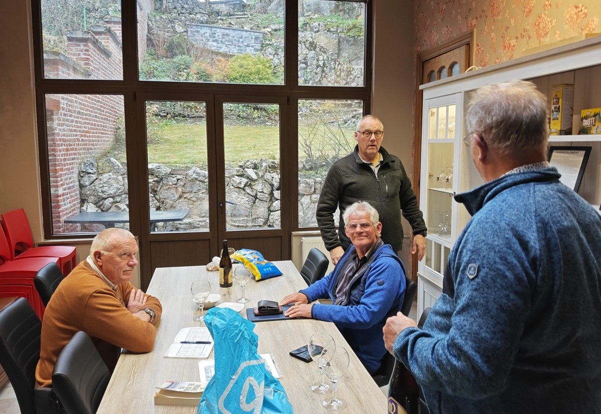 Vier mensen zitten en staan rond een houten tafel in een kamer met een groot raam.