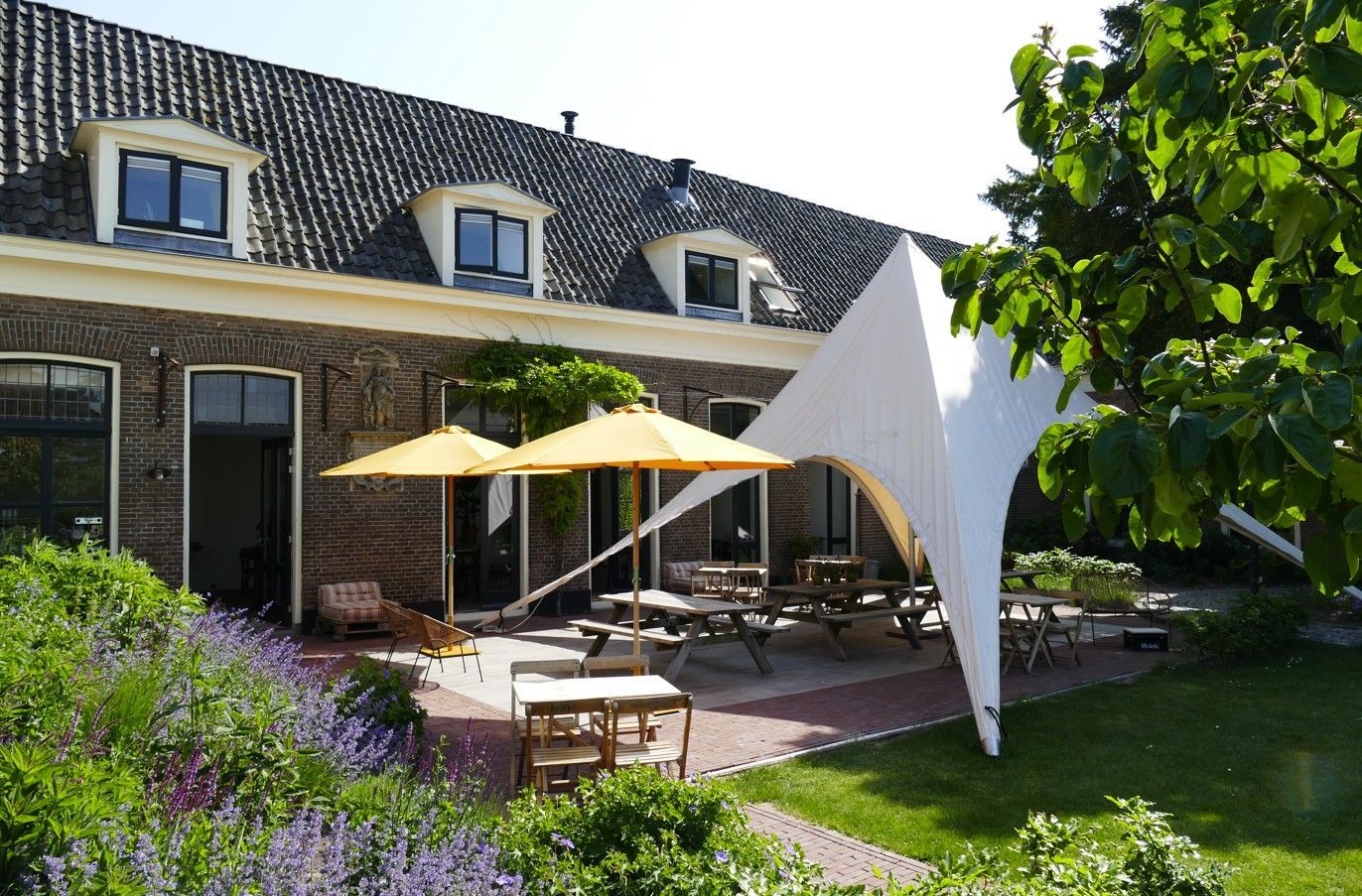 Buitenterras met tafels, parasols en een bijzondere witte tent naast een bakstenen gebouw.