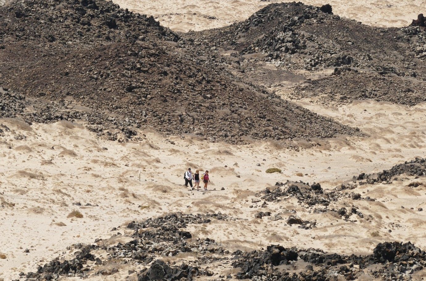 Wandelaars lopen door een zanderig, rotsachtig woestijnlandschap. Bruin en beige overheersen.