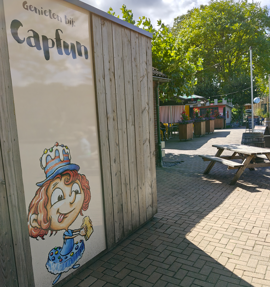 Cafékiosk met muurschildering van een stripfiguur, houten gevelbekleding en picknicktafels buiten op een zonnig plein.