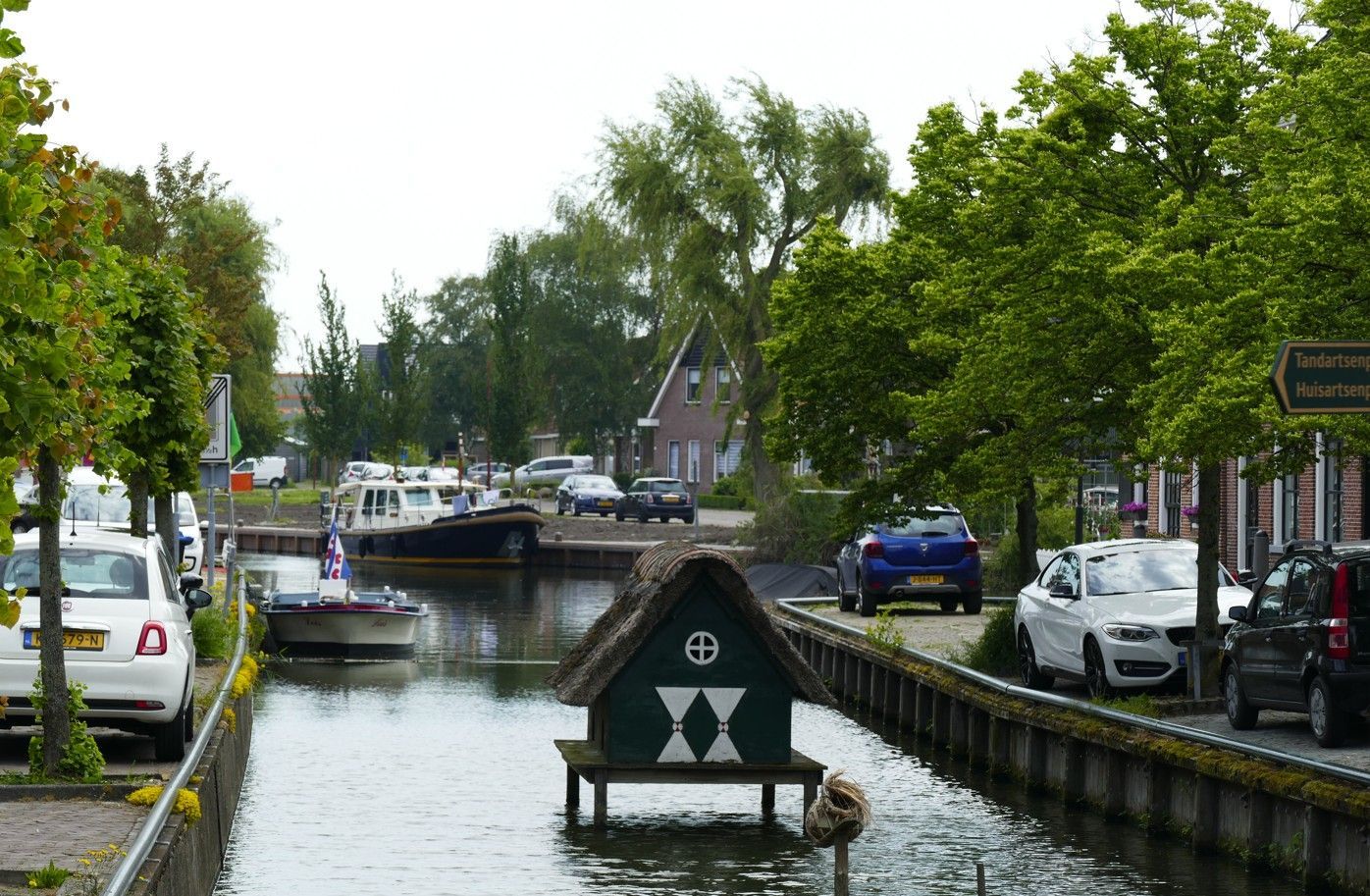 Een kanaal omzoomd met bomen en geparkeerde auto's, een boot en een klein gebouwtje met een rieten dak.
