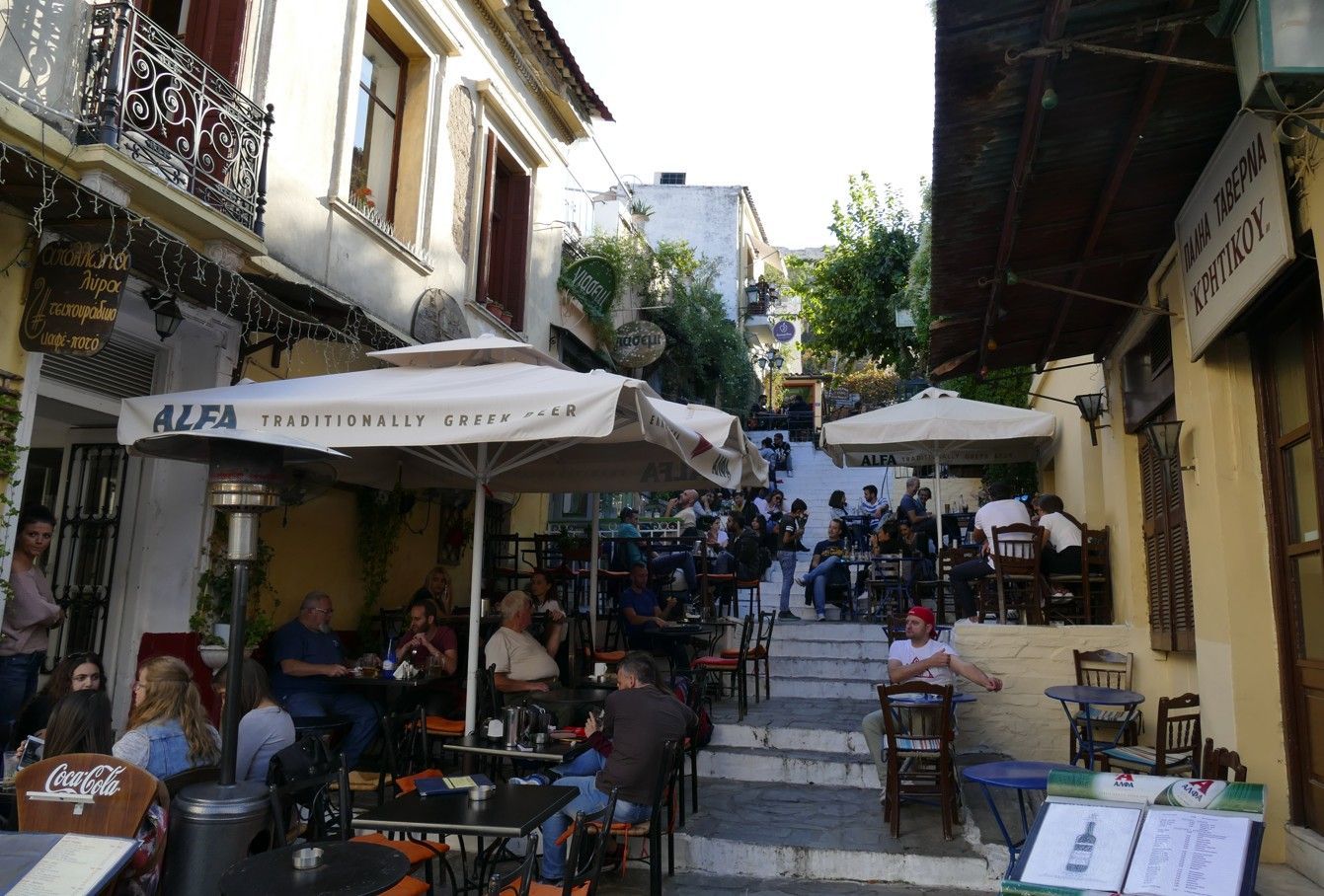 Een terrasje in een smalle straat met trappen. Mensen zitten aan tafels onder parasols, anderen beklimmen de trap.