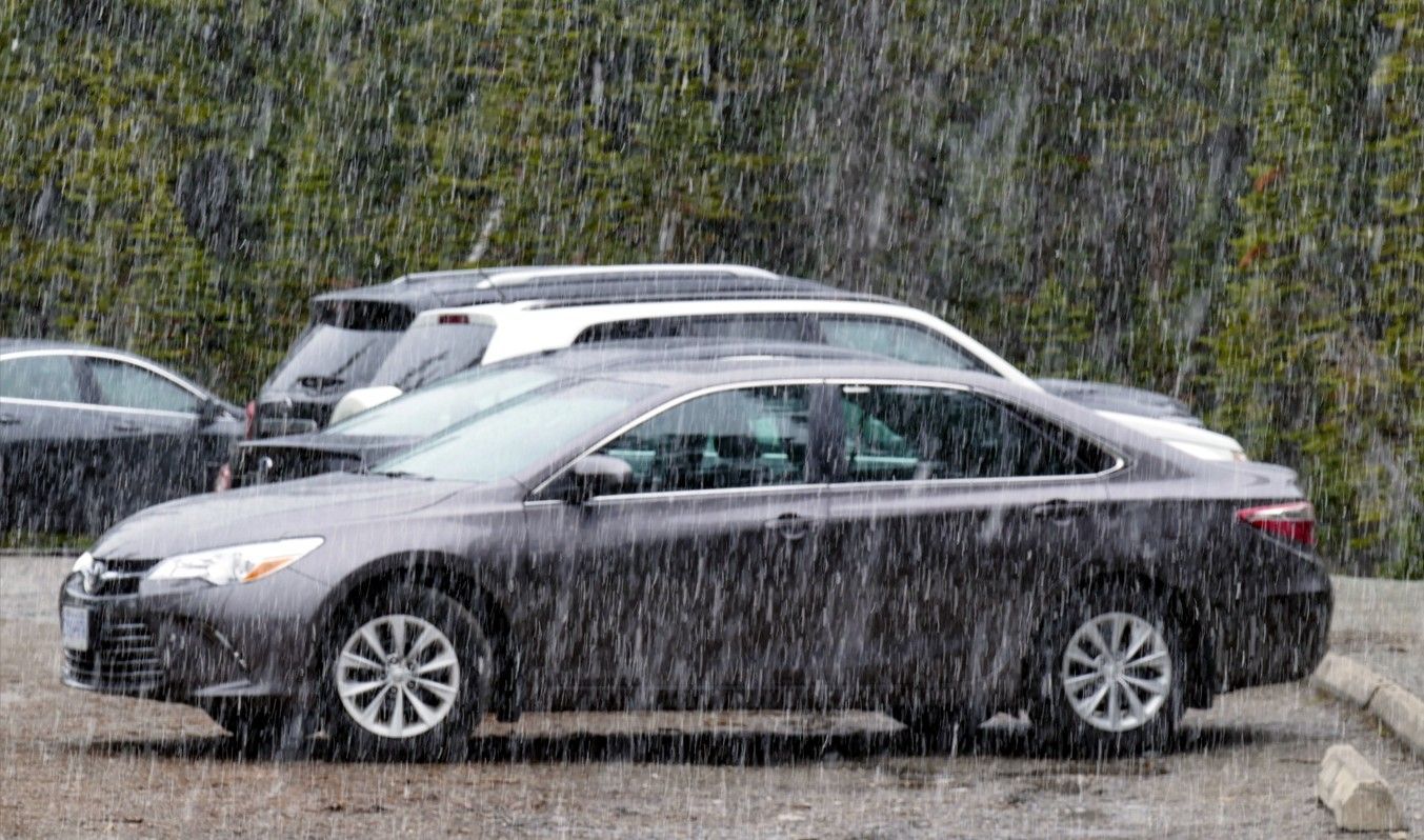 Donkergrijze auto geparkeerd in de regen, met andere auto's en bomen op de achtergrond.