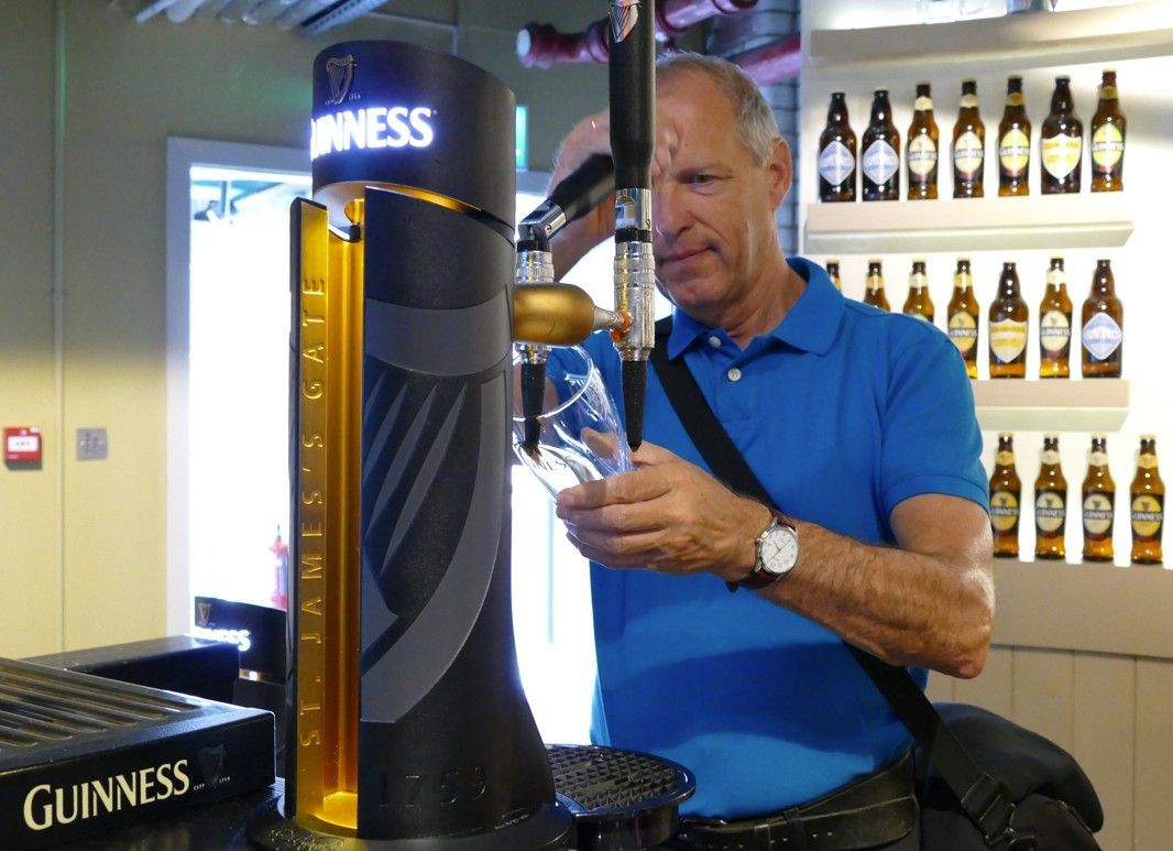 Jan tapt Guinness uit een tapkraan. Hij leert Guiness tappen. Op de achtergrond zijn bierflesjes te zien.