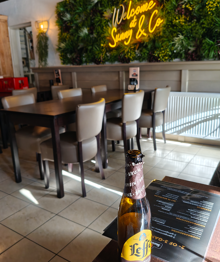 Gezellig restaurantinterieur met houten tafels, planten aan de muur, een neonreclame en een flesje bier op tafel.