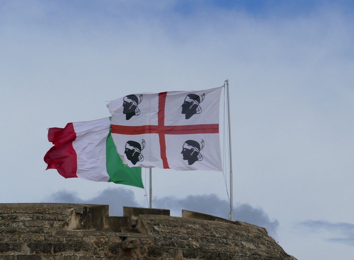 De Italiaanse en Sardijnse vlaggen wapperen tegen een bewolkte hemel op een stenen constructie.