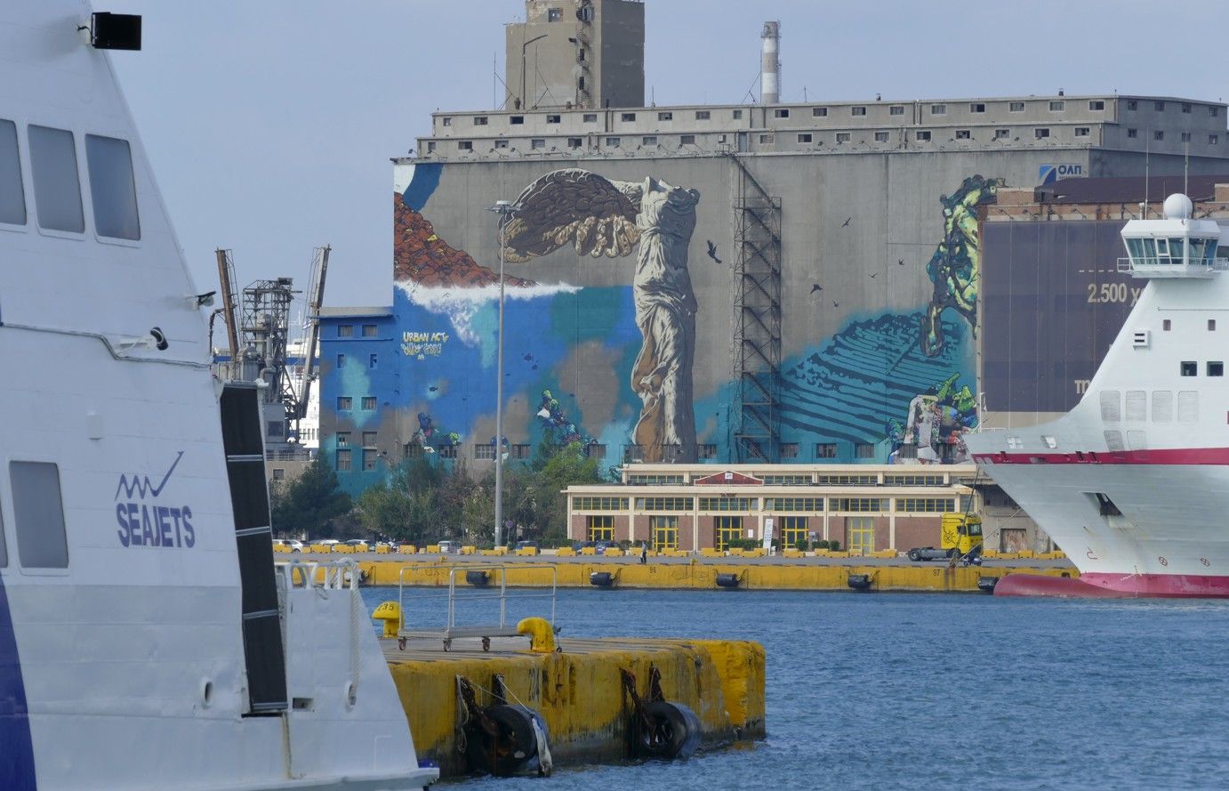 Twee veerboten op het water, met op de achtergrond een industrieel gebouw bedekt met muurschilderingen en een gele aanlegsteiger.