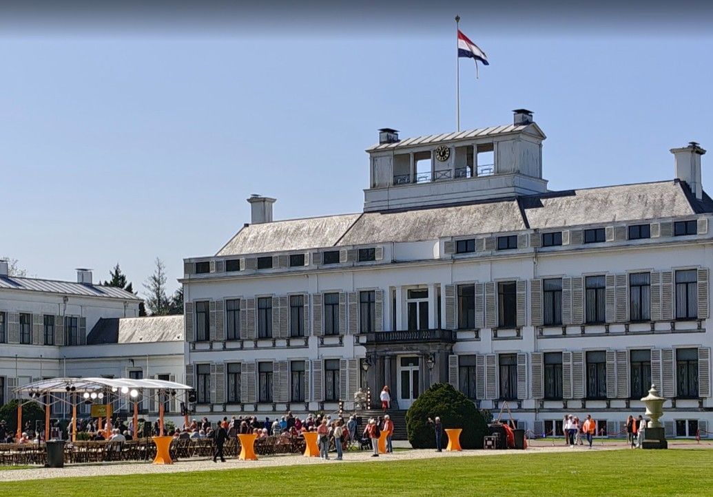 Paleis met Nederlandse vlag, mensen op het gazon, oranje versieringen en een helderblauwe lucht.