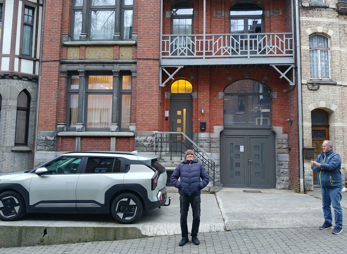 Twee mensen staan op een straat met kinderkopjes voor een bakstenen gebouw, met een auto ernaast geparkeerd.