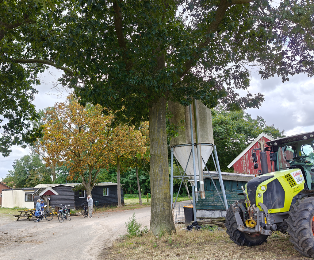 Een landelijk gelegen boerderij met een tractor, silo, schuur, bomen en een persoon in de buurt.