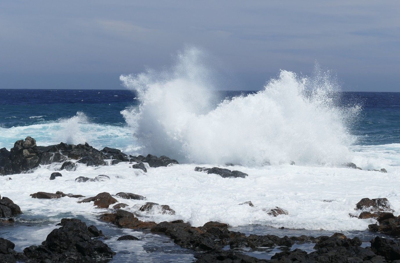 Golven beuken tegen zwarte rotsen en vormen een grote waternevel tegen een blauwe lucht en oceaan.