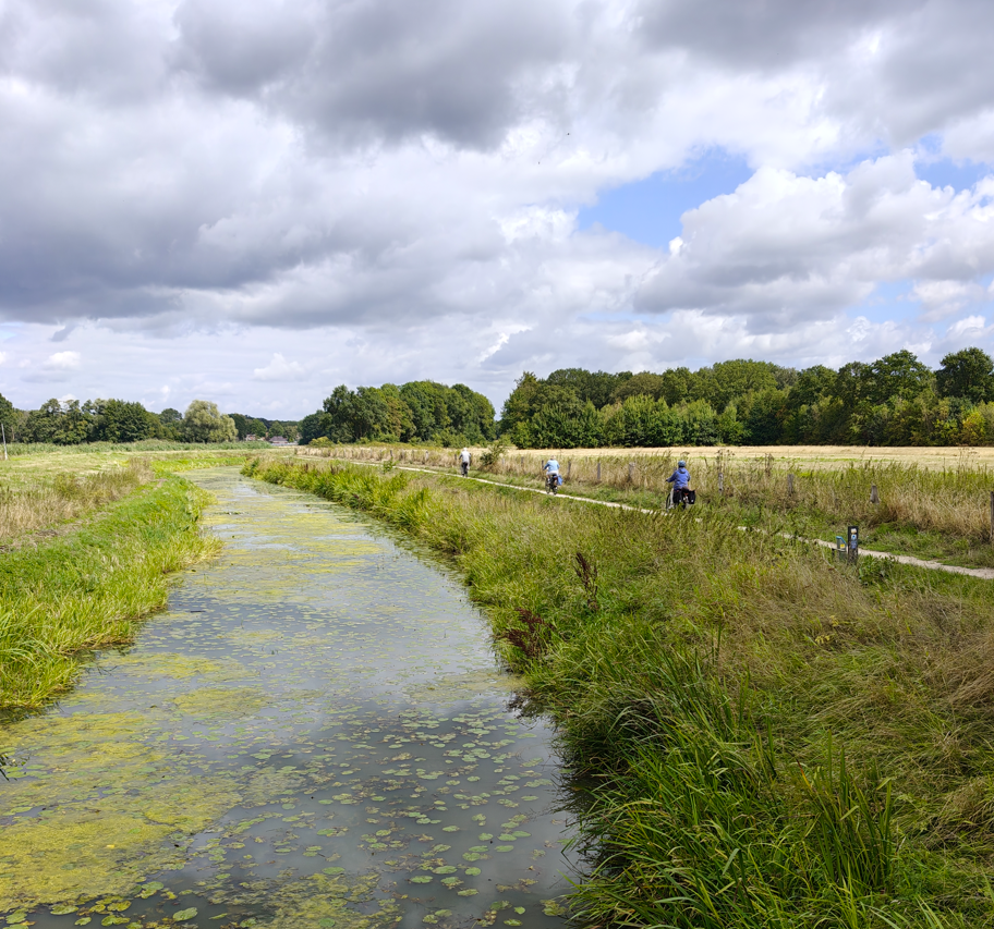 Een smal kanaal, omzoomd met gras en wilde bloemen, onder een bewolkte hemel, met een paar mensen die over het pad wandelen.