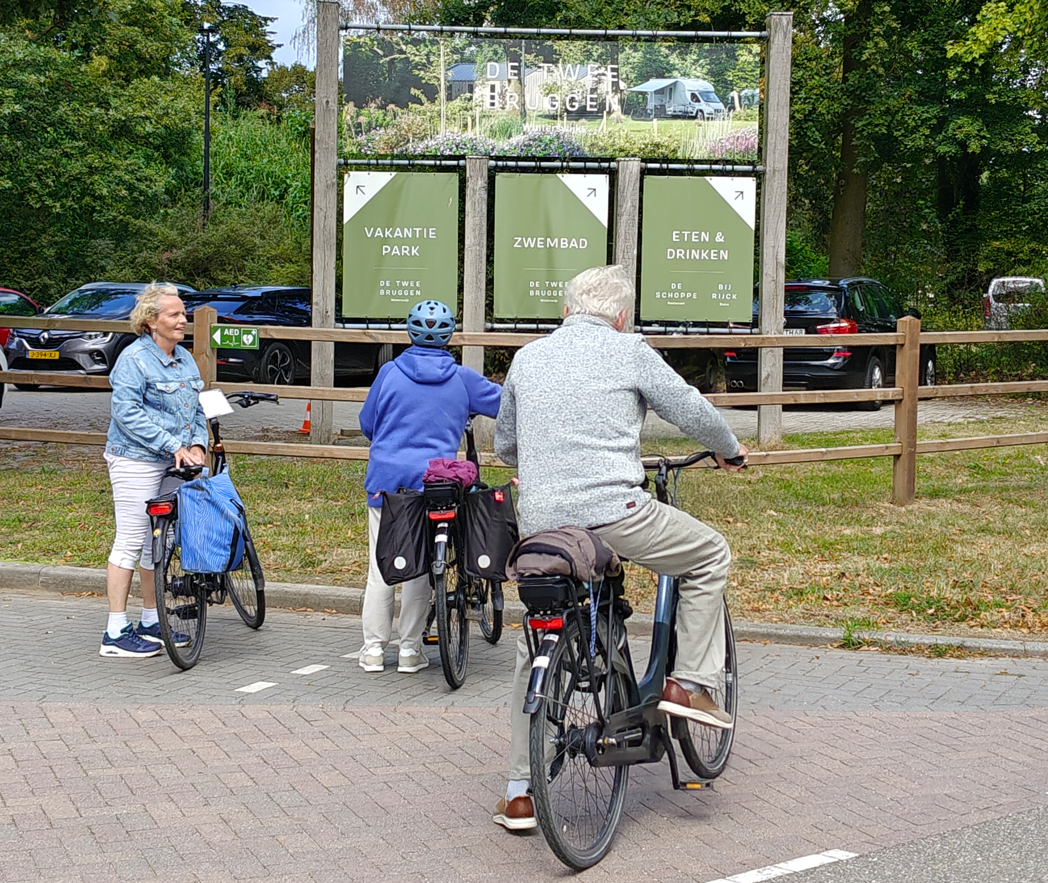 Fietsers en een voetganger verzamelden zich bij een bord van een parkpad aan de ingang van een bebost gebied langs de weg.