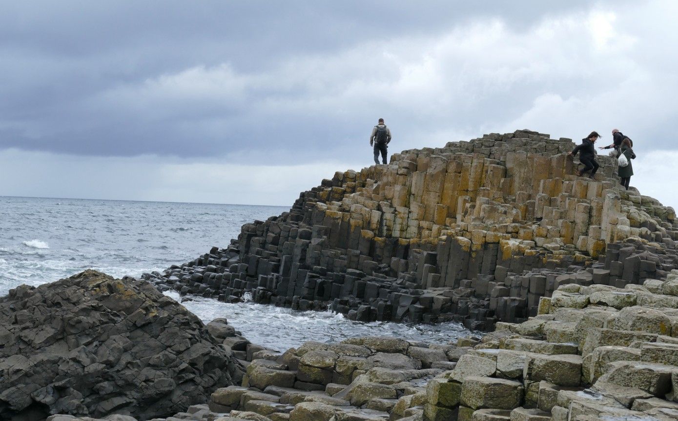 Mensen op zeshoekige basaltkolommen bij de Giant's Causeway in Noord-Ierland, aan zee onder een bewolkte hemel.