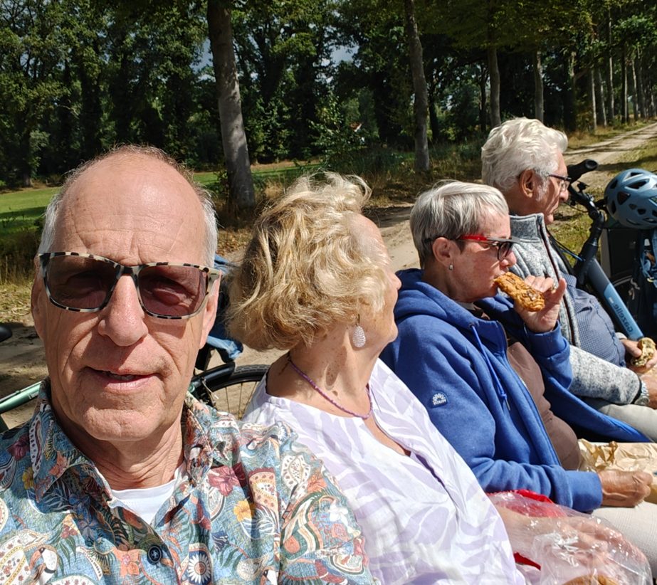 Mensen zitten op een bankje in een zonnig park, met een man met een zonnebril op de voorgrond.