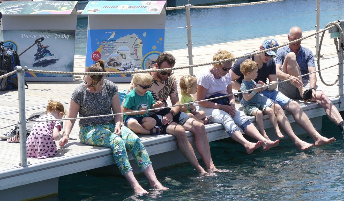 Mensen zitten met hun voeten in het water op een steiger. Achter hen hangen borden en er is een touwreling aanwezig.