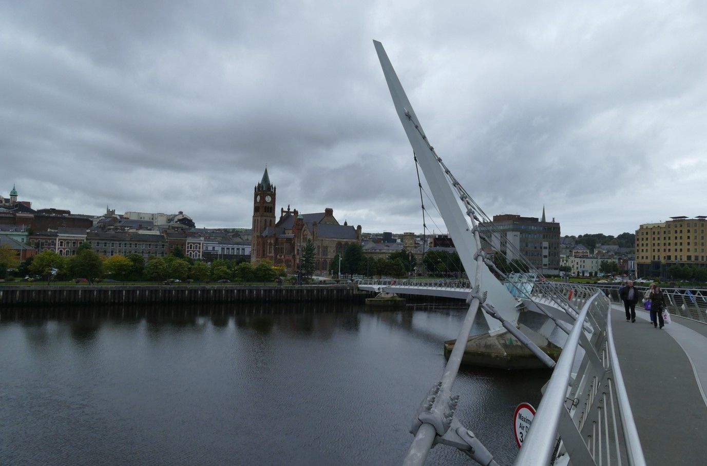 Moderne brug met centrale spits over een rivier, die onder een bewolkte hemel stadsgebouwen met elkaar verbindt.