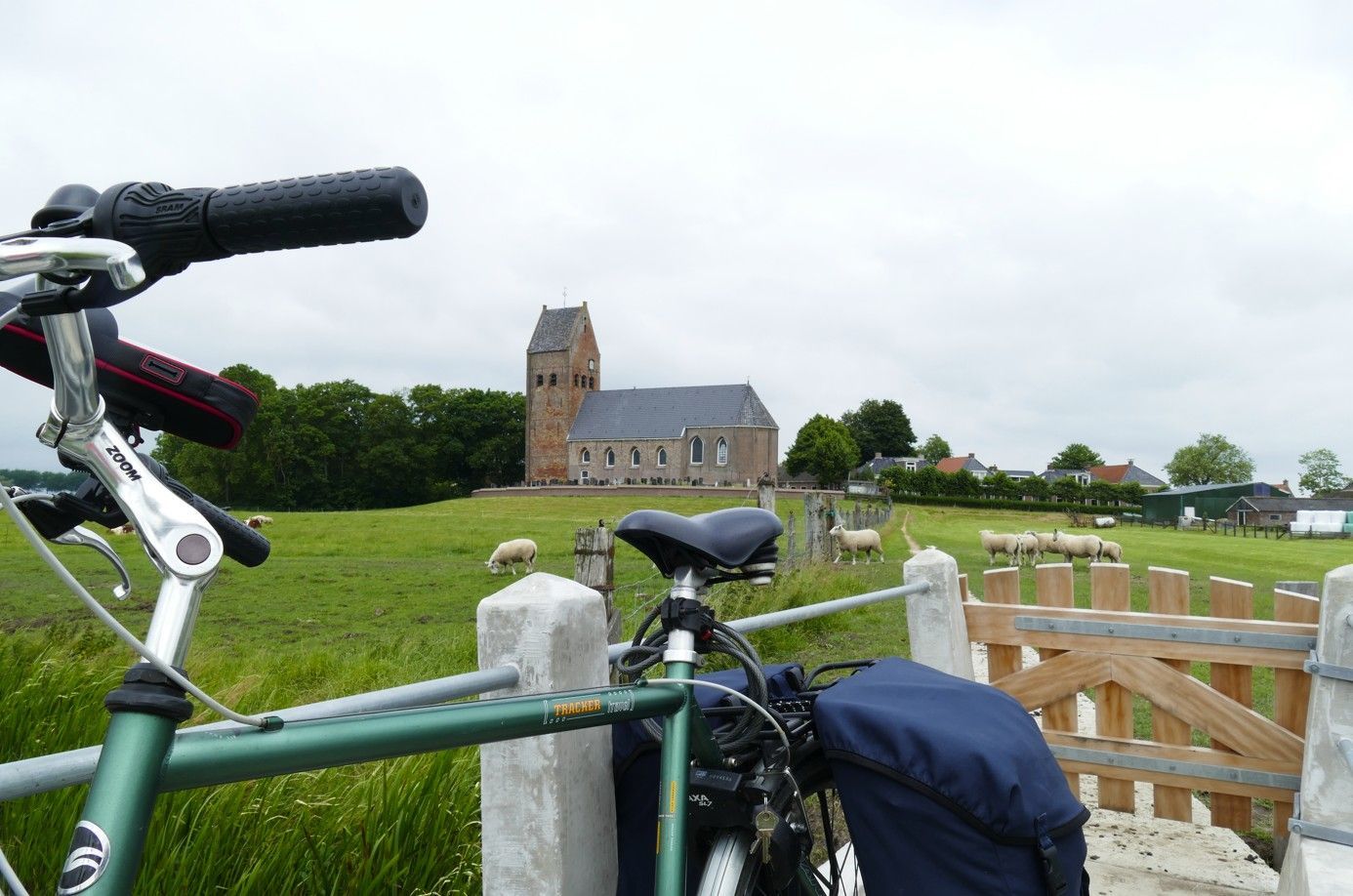 Fiets staat geparkeerd bij een hek, met op de achtergrond een kerk op een bewolkte dag. Schapen grazen in een veld.
