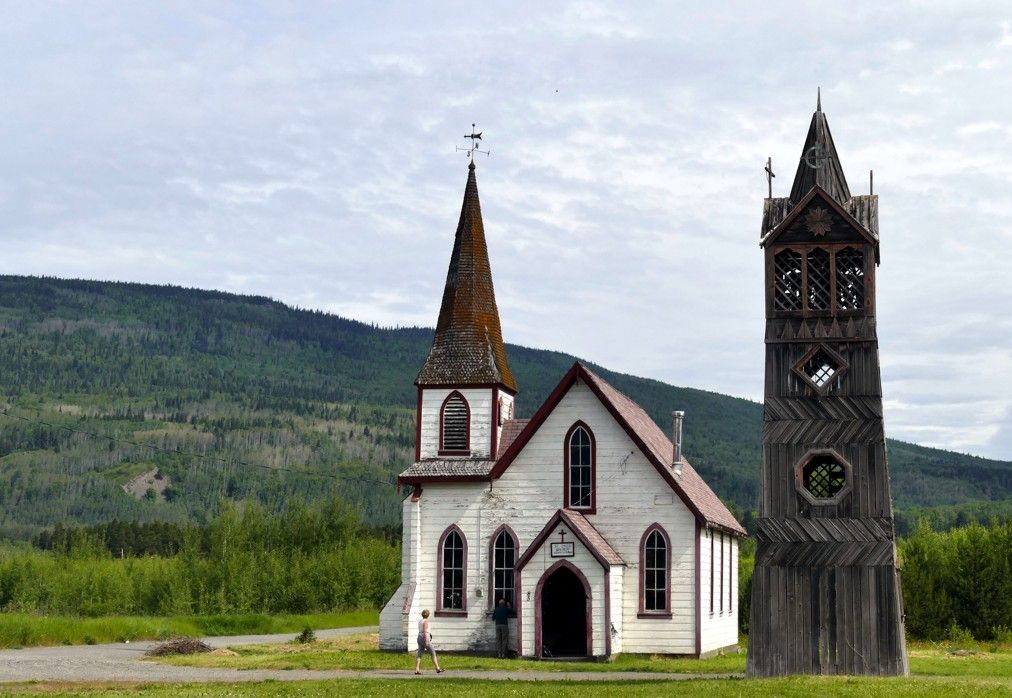 Een witte houten kerk met klokkentoren staat in een veld met bergen op de achtergrond, onder een bewolkte hemel.