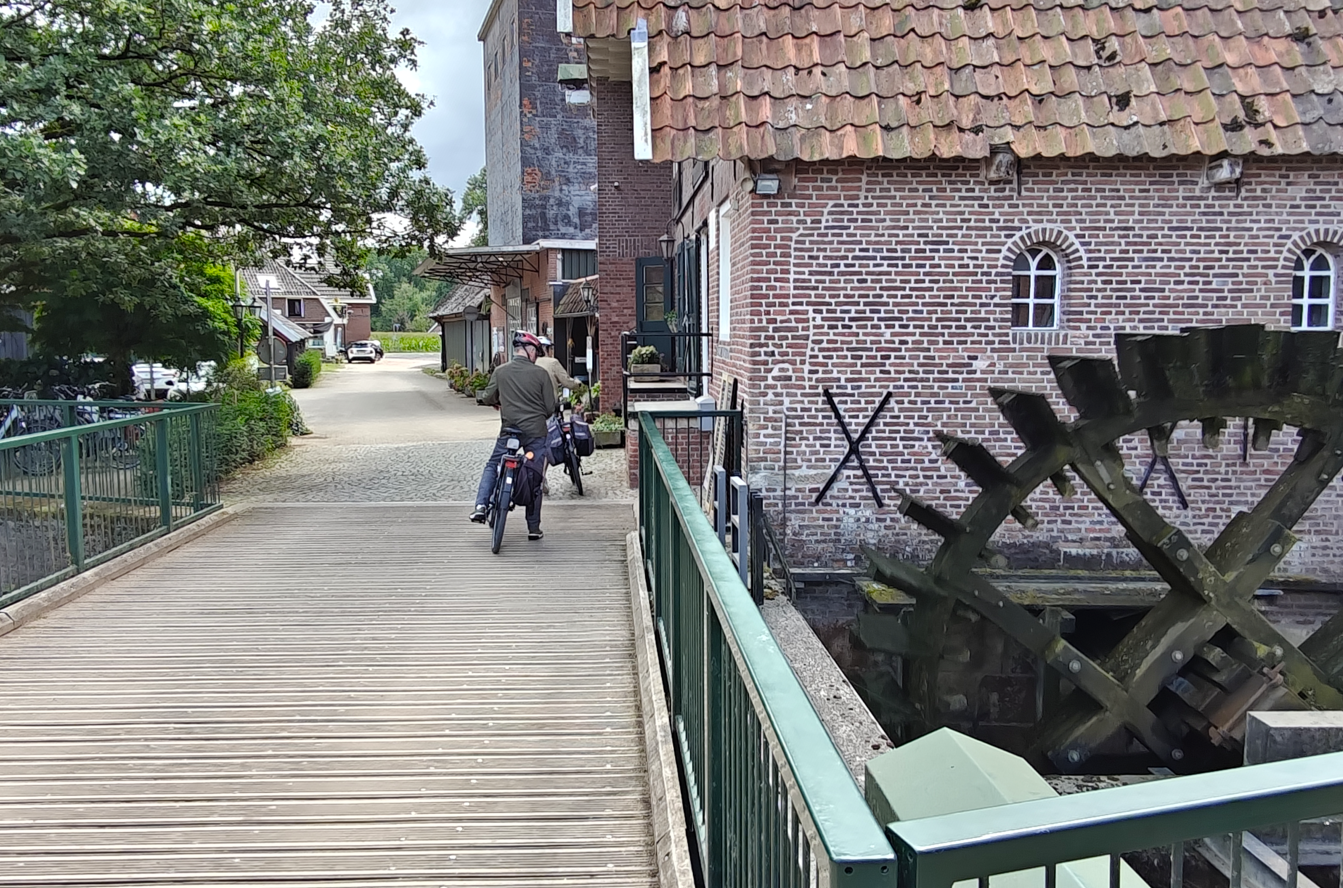 Houten vlonderpad naast een bakstenen watermolen, met een groot waterrad en twee mensen die vooruit lopen.