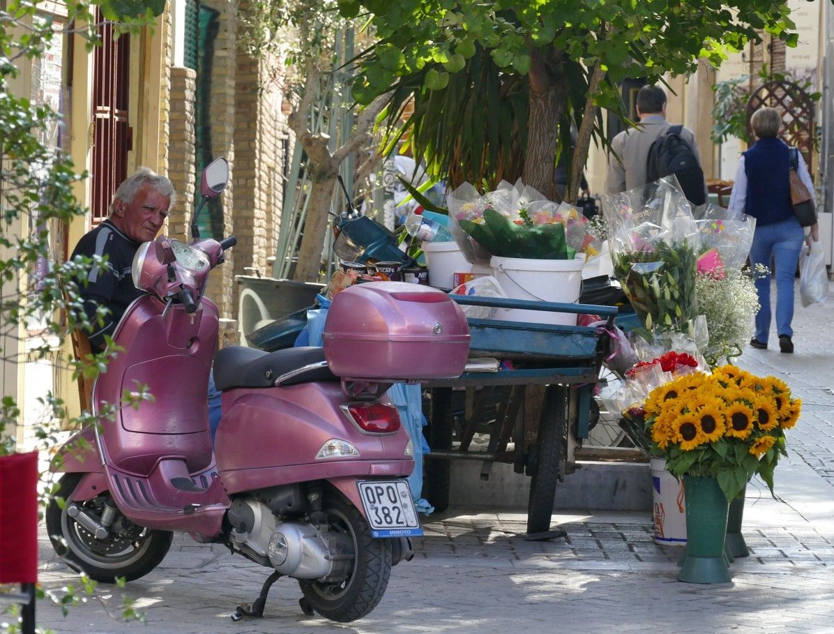 Paarse scooter geparkeerd bij een bloemenkraam in een Europese straat, verkoper ernaast, voetgangers op de achtergrond.