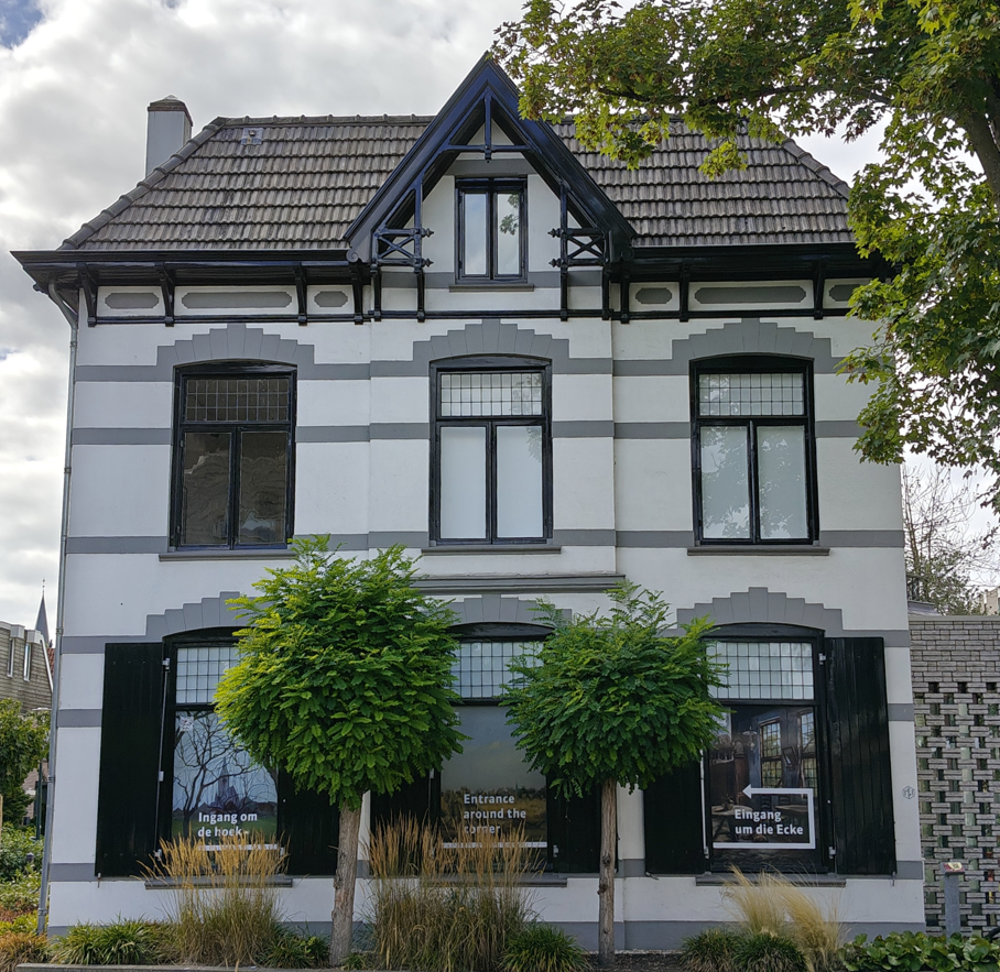 Zwart-wit huis met twee verdiepingen, een zadeldak en gesnoeide bomen aan de voorzijde.