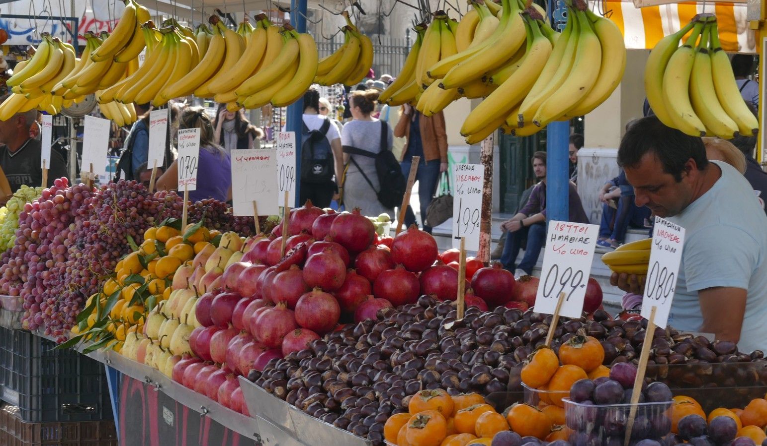 Fruitkraam met bananen, druiven, granaatappels en een verkoper op een openluchtmarkt.