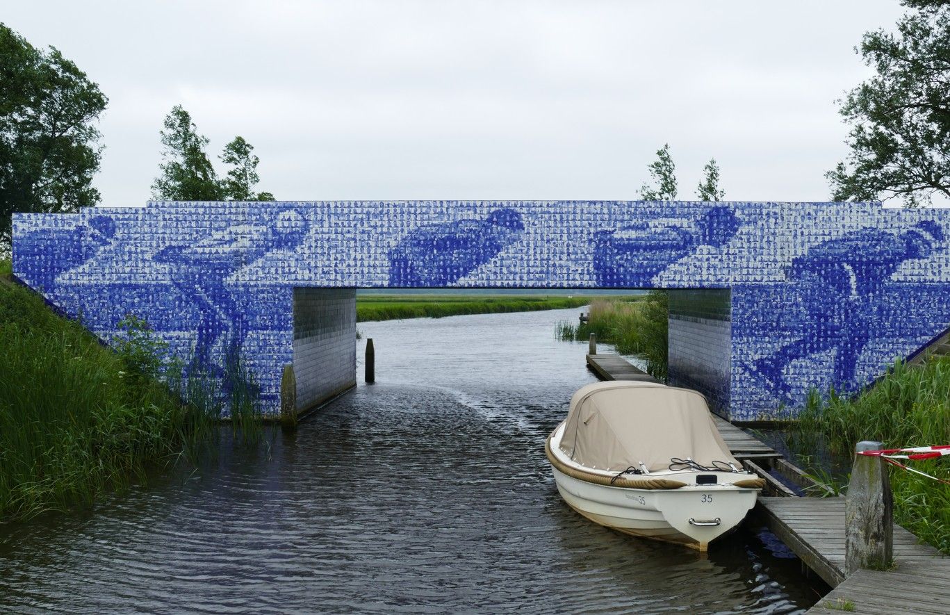 Een brug, versierd met blauwe mozaïeken van schaatsers, overspant een kanaal; een boot ligt eronder aangemeerd.