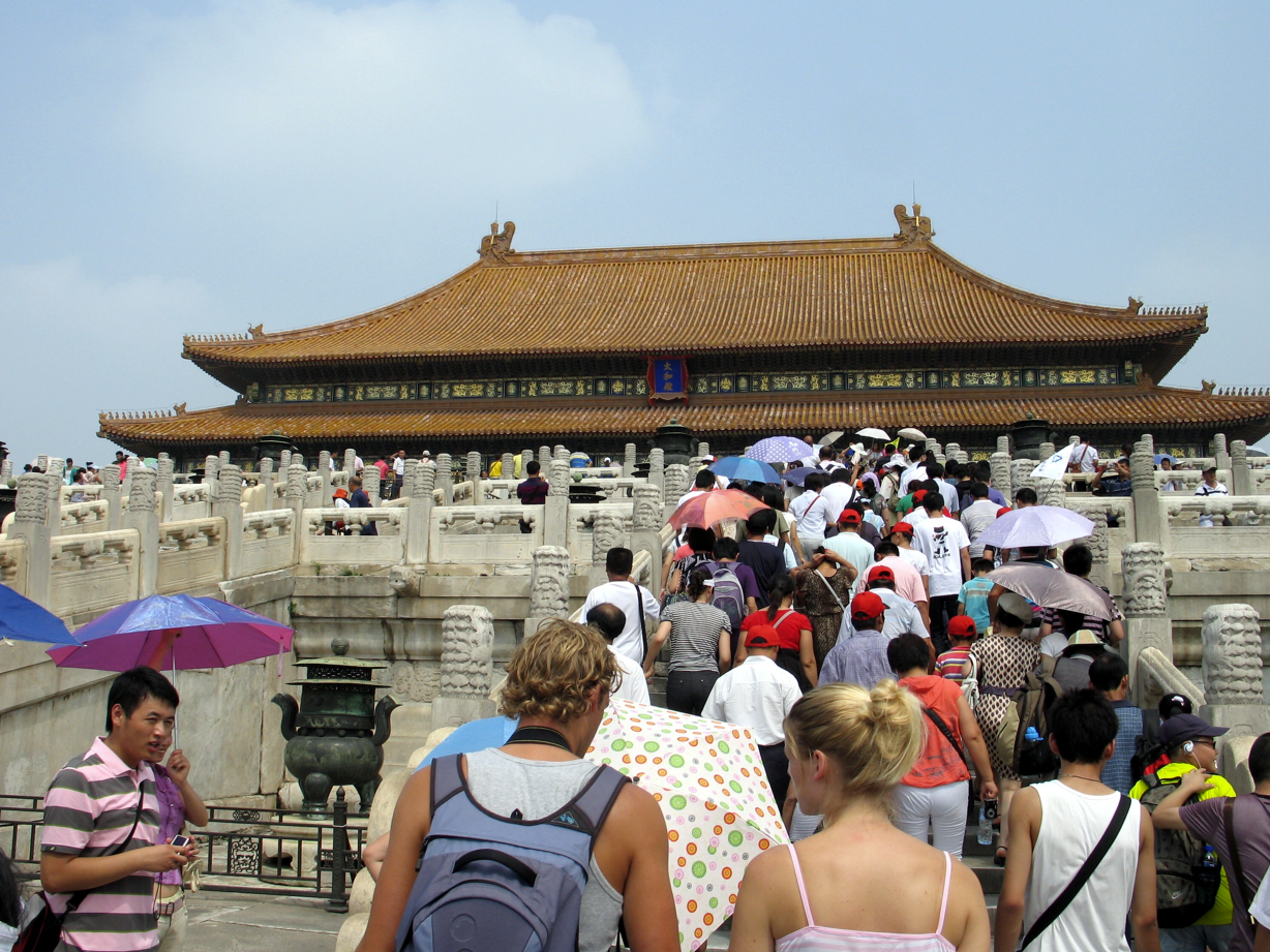 Mensen beklimmen trappen naar een Chinees paleis, velen met een paraplu in de hand tegen de zon.