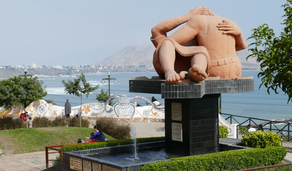 Standbeeld van omhelzende geliefden aan zee, met een fontein ervoor en een kuststad op de achtergrond.